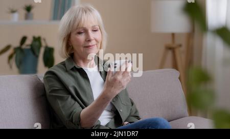 Vieille femme caucasienne 60s la femme âgée mûre est assise à la maison sur un canapé avec le téléphone portable lit les nouvelles avis pense que les rêves réfléchissent l'idée de regarder loin Banque D'Images