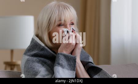 Gros plan malade vieille grand-mère blonde 60s femme couverte d'une couverture chaude à la maison nez soufflant à la serviette en papier souffre nez courant souffrent de Banque D'Images