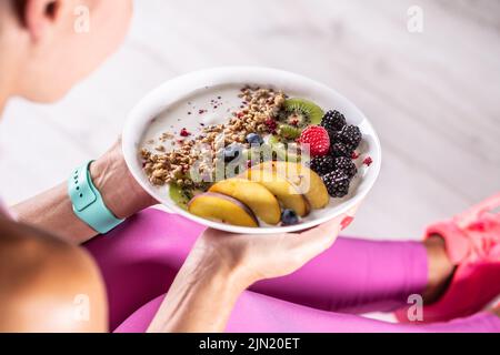 Une femme a un petit déjeuner sain après l'exercice du matin. Yaourt, mûres muesli, framboises, bleuets, kiwis et pêches dans un bol blanc. Banque D'Images