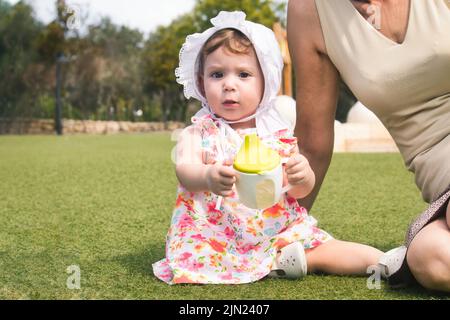 Une petite fille adorable, portant un bonnet blanc et une robe, assise sur l'herbe verte avec sa grand-mère et tenant une tasse à bec avec de l'eau Banque D'Images