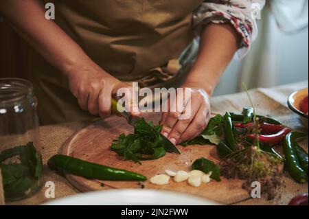 Gros plan de la femme de ménage hachant des herbes fraîches sur une planche de bois tout en préparant la saumure pour le pickling à la cuisine maison Banque D'Images