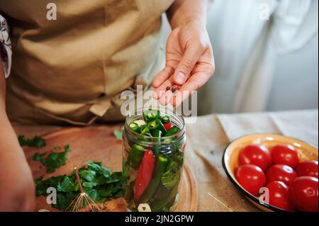 Femme à la main ajoutant des grains de poivre dans des jarres de verre avec des piments frais empilés tout en faisant des conserves à la cuisine maison Banque D'Images