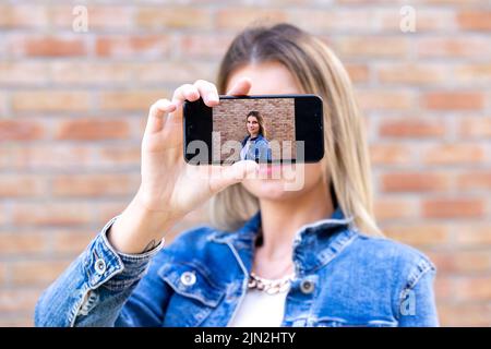 Photo de gai mignon charmant joli joli joli joli joli joli joli joli petit-fille blond, pâle peau montrant selfie avec son téléphone tenu dans ses mains portant un Jean veste isolée sur fond de brique de couleur rouge. Photo de haute qualité Banque D'Images