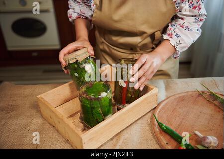 Les mains de la femme de ménage mettant des bocaux en conserve avec des concombres marinés et des piments marinés à l'envers sur une caisse en bois Banque D'Images