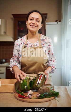 Charmante femme de ménage sourira en montrant des bocaux avec des concombres marinés et des piments. Mise en conserve saisonnière de légumes Banque D'Images