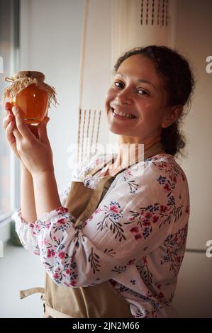 Belle femme afro-américaine, portant un tablier beige tient un pot avec des confiture de pêche et des sourires regardant la caméra Banque D'Images