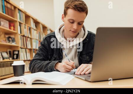 Un étudiant de sexe masculin écrit tout en étant assis à son bureau dans la bibliothèque de l'université Banque D'Images