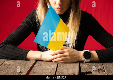 Une fille s'assoit à une table à la maison et tient le drapeau national de l'Ukraine sur fond d'un mur rouge, arrêter la guerre en Ukraine, je suis ukrainien 2022 Banque D'Images