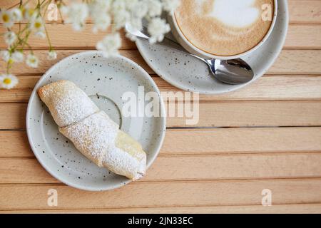 Croissant français maison avec poudre. Tasse de cappuccino en céramique blanche, avant-plan flou de fleurs de camomille sur une table en bois au Banque D'Images