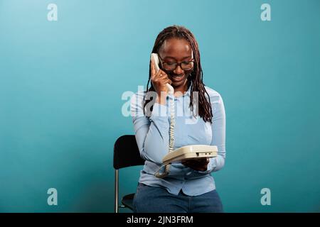 Un opérateur de téléphone fixe souriant ayant une conversation sur un téléphone filaire sur fond bleu. Jeune femme adulte confiante et positive répondant aux appels des clients pour des services de conseil. Banque D'Images