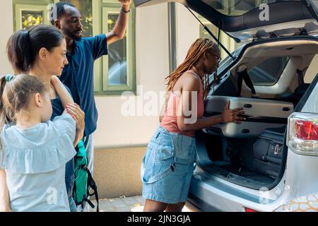 Divers membres de la famille et amis partent en voyage, chargeant des sacs gonflables et des sacs à roulettes dans le coffre du véhicule pour voyager pendant les vacances d'été. Aller à la destination balnéaire avec des gens souriants. Banque D'Images