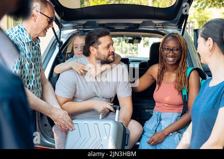 Divers membres de la famille et des amis chargent le chariot dans le coffre d'automobile, se préparant à partir en vacances. Voyage en voiture pendant les vacances d'été, aller à destination de bord de mer avec des personnes multiethniques. Banque D'Images