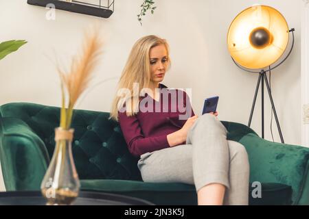 Jeune femme blonde caucasienne cherchant focalisée à la recherche de quelque chose sur le smartphone assis sur le canapé dans la salle de séjour. Prise de vue en intérieur. Photo de haute qualité Banque D'Images