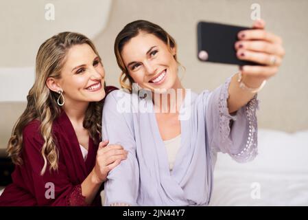 Les jours enfin ici. Une belle jeune mariée et sa meilleure amie prenant un selfie ensemble dans leur dressing avant le mariage. Banque D'Images