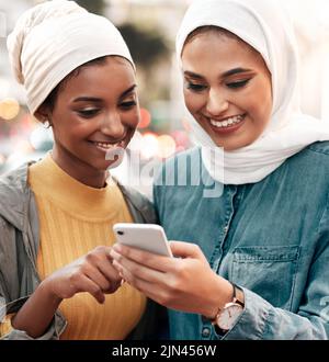 Allons voir cet endroit. Deux jeunes femmes attrayantes portant un foulard et debout ensemble tout en utilisant un téléphone cellulaire dans la ville. Banque D'Images