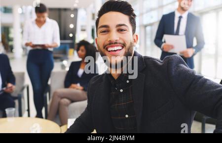 Je me sourit de relever des défis. Portrait court d'un beau jeune homme d'affaires assis et souriant tandis que ses collègues travaillent derrière lui dans le Banque D'Images