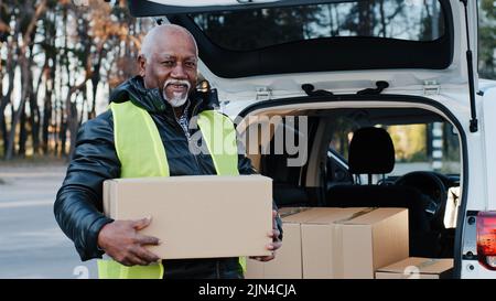 Mature afro-américain souriant homme tenant des supports de boîte en carton près du coffre ouvert de l'automobile heureux homme âgé courier dehors dans les vêtements de travail Banque D'Images