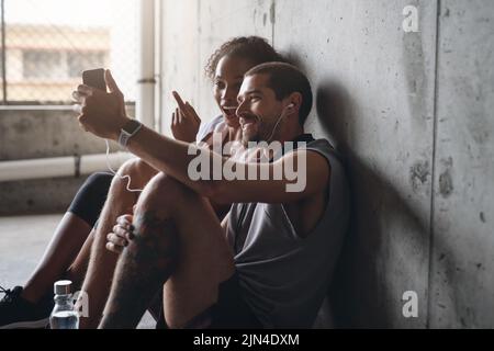 Créer l'ambiance pour notre prochain entraînement. Un jeune couple sportif utilisant un téléphone portable ensemble tout en étant assis et en se reposant contre un mur. Banque D'Images