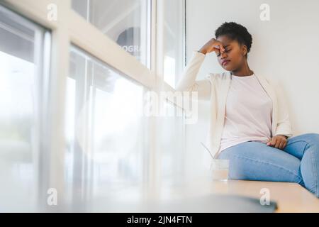 Femme afro-américaine assise sur le seuil de la fenêtre fatiguée, frottant le nez et les yeux sentant la fatigue. Dépression et tristesse. Contrarié et irrité pour le problème Banque D'Images