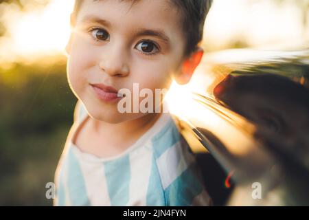 Gros plan d'un garçon qui tient sa tête par la fenêtre de la voiture et regarde l'appareil photo. Voyagez en toute liberté. Bonne famille, enfance. Voyage en voiture. Banque D'Images