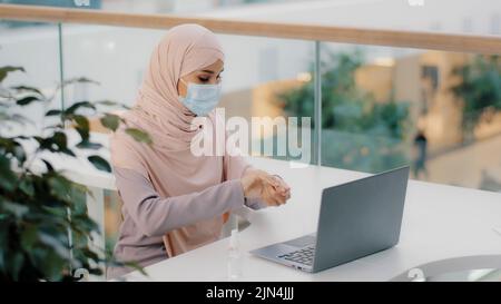 Drôle de femme arabe assis dans un lieu public utilise des arrosettes antiseptiques autour dans l'air sur ordinateur couvre le visage avec masque médical fille musulmane peur des germes Banque D'Images