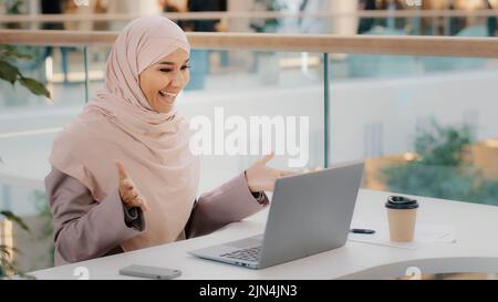 Bonne jeune femme d'affaires arabe assise au bureau, dactylographiant sur un ordinateur portable qui travaille sur Internet, vérifiant une nouvelle application souriant en regardant l'appareil photo avec le pouce vers le haut Banque D'Images