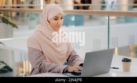 Bonne jeune femme d'affaires arabe assise au bureau, dactylographiant sur un ordinateur portable qui travaille sur Internet, vérifiant une nouvelle application souriant en regardant l'appareil photo avec le pouce vers le haut Banque D'Images