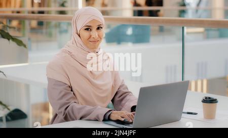 Bonne jeune femme d'affaires arabe assise au bureau, dactylographiant sur un ordinateur portable qui travaille sur Internet, vérifiant une nouvelle application souriant en regardant l'appareil photo avec le pouce vers le haut Banque D'Images