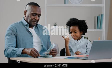 Joyeux père d'adulte et petite fille mignonne assis à la maison papa enseigne enfant fille à compter montre des dollars en espèces des factures de parent de soin aide l'enfant à apprendre Banque D'Images