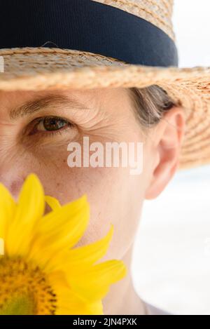 Portrait court de la tête d'une femme aux cheveux verts gris en chapeau de paille couvrant la partie inférieure de son visage avec du tournesol, regardant attentivement l'appareil photo. Banque D'Images