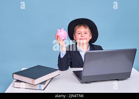 Un garçon dans un costume d'école avec une banque de piggy et un chapeau noir à un ordinateur pendant l'apprentissage à distance, un espace de copie sur un fond bleu studio Banque D'Images