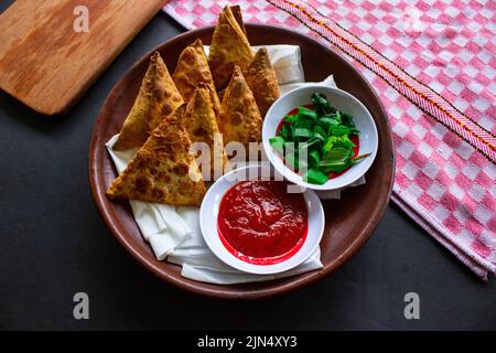 Samosa, samsa ou somsa sont des pâtisseries frites triangulaires ou pastri servies avec une sauce tomate et une tranche de poireaux, sur fond noir Banque D'Images