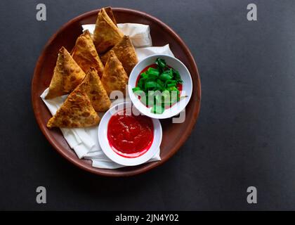 Samosa, samsa ou somsa sont des pâtisseries frites triangulaires ou pastri servies avec une sauce tomate et une tranche de poireaux, sur fond noir Banque D'Images