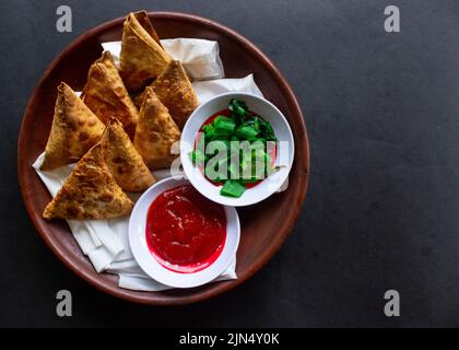 Samosa, samsa ou somsa sont des pâtisseries frites triangulaires ou pastri servies avec une sauce tomate et une tranche de poireaux, sur fond noir Banque D'Images