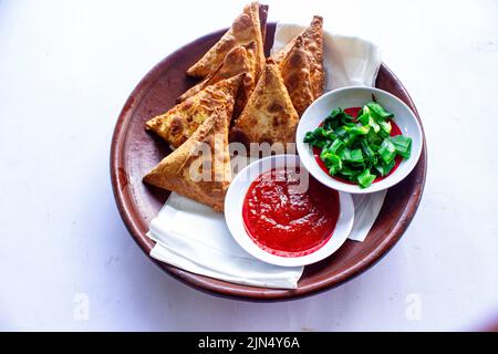 Samosa, samsa ou somsa sont des pâtisseries frites triangulaires ou pastri servies avec une sauce tomate et une tranche de poireaux, sur fond noir Banque D'Images