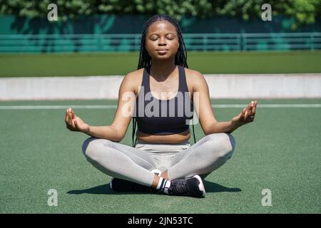 Une femme concentrée médite assise à Lotus poser sur un terrain vert au stade contre les arbres près du parc de la ville Banque D'Images
