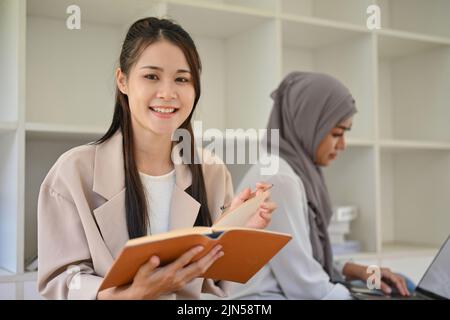 Une jeune étudiante asiatique attrayante lit un livre dans l'espace de travail conjoint de la bibliothèque du campus. Banque D'Images