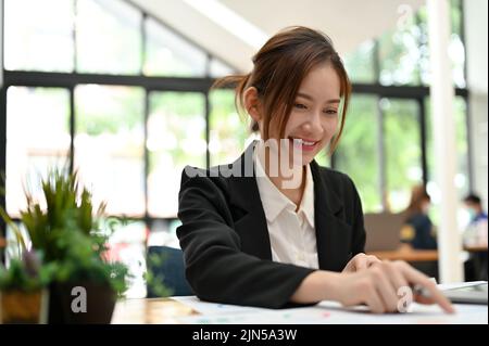 Une jolie jeune femme d'affaires asiatique se trouve dans l'espace de travail de son bureau et travaille sur la paperasse de son rapport financier. Banque D'Images