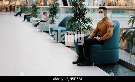 Jeune indien mec dans un masque médical protégeant la santé pendant une pandémie assis dans un fauteuil à l'intérieur dans le centre commercial repos relaxant nerveux Banque D'Images