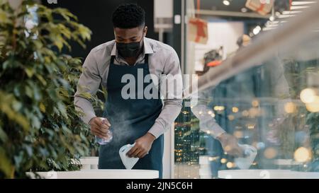 Jeune homme serveur dans le tablier lingettes vaporisateurs de table nettoyant désinfectant prépare le café restaurant pour l'ouverture d'un étudiant afro-américain sérieux en médecine Banque D'Images