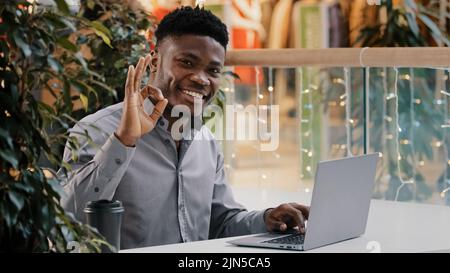 Homme d'affaires confiant et concentré travaillant à distance en tapant sur un ordinateur portable jeune homme sérieux regardant la caméra sourire montrant le signe ok approuvant le geste Banque D'Images