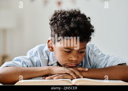 Un garçon africain sérieux se concentrant sur la lecture d'un livre à table à la maison Banque D'Images