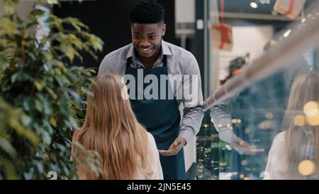 Un jeune serveur sympathique souriant servi en tablier sert une cliente dans un café. Un barman, un ouvrier du restaurant, apporte une boisson au café dans une tasse de papier Banque D'Images