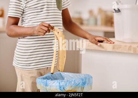 Gros plan d'un garçon africain qui jette le zeste de banane dans un seau tout en se tenant dans la cuisine Banque D'Images