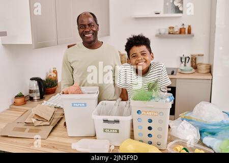 Portrait d'un heureux papa africain triant les ordures dans des conteneurs séparés avec son fils dans la cuisine Banque D'Images