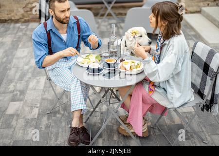 Un homme et une femme élégants ont un petit déjeuner tout en étant assis avec un chien dans un café à l'extérieur Banque D'Images