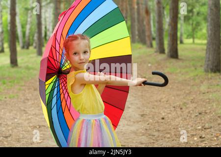 Bonne fille drôle tenant parapluie arc-en-ciel. Adorable écolière joyeuse jouant dans un parc d'été pluvieux. Enfant marchant dans la douche d'automne. Des activités de plein air Banque D'Images