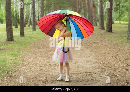 Bonne fille drôle tenant parapluie arc-en-ciel. Adorable écolière joyeuse jouant dans un parc d'été pluvieux. Enfant marchant dans la douche d'automne. Des activités de plein air Banque D'Images