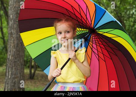 Bonne fille drôle tenant parapluie arc-en-ciel. Adorable écolière joyeuse jouant dans un parc d'été pluvieux. Enfant marchant dans la douche d'automne. Des activités de plein air Banque D'Images