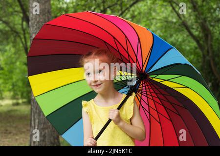 Bonne fille drôle tenant parapluie arc-en-ciel. Adorable écolière joyeuse jouant dans un parc d'été pluvieux. Enfant marchant dans la douche d'automne. Des activités de plein air Banque D'Images